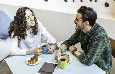 Smiling young man and woman talking over coffee and croissants at a cafe table with a tablet. Concept of dating, friendship, modern lifestyle and relaxed breakfast moments.