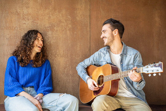 Young man playing acoustic guitar for a smiling friend outdoors. Concept of friendship, music, creativity, leisure and joyful moments together in an urban lifestyle setting. - Powered by Adobe