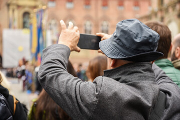 Tourist taking photo with smartphone during city event in Italy. Senior man in bucket hat capturing urban scene among a crowd. Travel lifestyle and street photography concept.