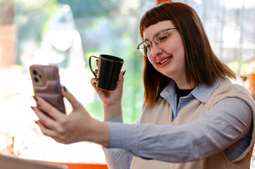 Young female employee or student taking a selfie with her mobile phone while drinking a coffee in the kitchen