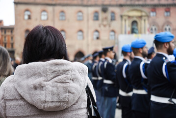 Woman attending a military ceremony in an Italian square, watching soldiers in uniform with blue berets. Rear view with blurred background of official event.