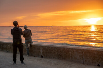 Asnelles-Sur-Mer, France - 08 08 2025: View of the jetty with a couple with dark dark clothes taking pictures with a smartphone of the sea and the sky at sunset