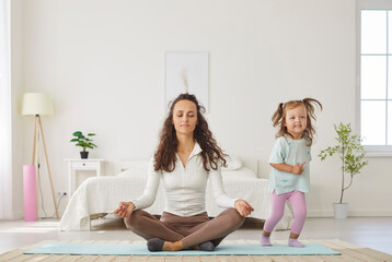 Relaxed mother sitting on floor in lotus pose, doing yoga breathing exercise with closed eyes and...