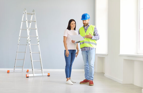 Male builder and construction engineer talking to female client. Man in yellow uniform vest and safety hat together with woman homeowner discuss indoor home remodeling and look at house plan on paper