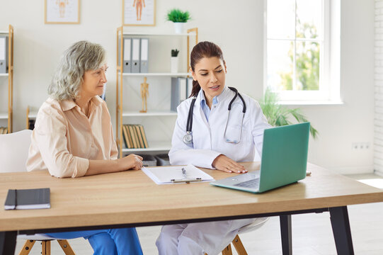 Cheerful friendly female doctor at medical office in hospital showing examination results on laptop monitor screen and giving consultation to a senior woman patient sitting in clinic.