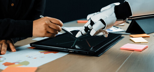 A human and a robot engage in a collaborative design process, using a touchscreen tablet to merge technology with creativity in a modern workspace.Noogenesis