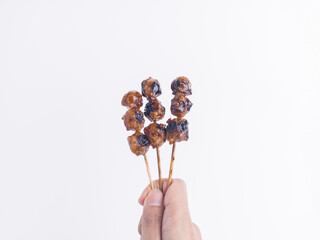 Hand holding three skewers of grilled Indonesian meatball satay with spicy peanut sauce, isolated...