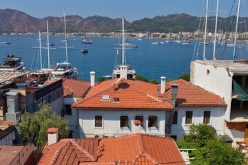 An aerial view of a white building with an orange tiled roof, set against a backdrop of a vibrant, sunny harbor. Sailboats dot the water in the distance in old Marmaris - Mugla - Turkey