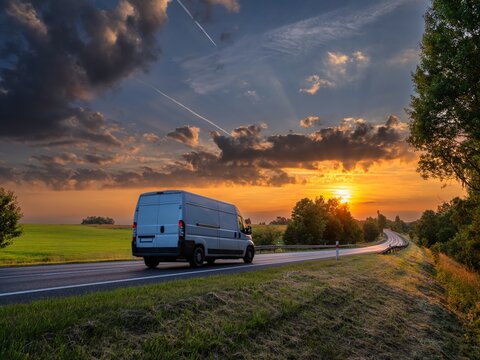 White fast delivery van driving on the asphalt road in rural landscape at golden sunset with dramatic clouds