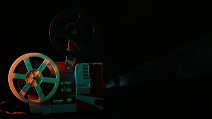 A vintage film projector with two reels, set against a black background. The projector is illuminated by colored lights, casting a beam of light into the darkness. - Powered by Adobe