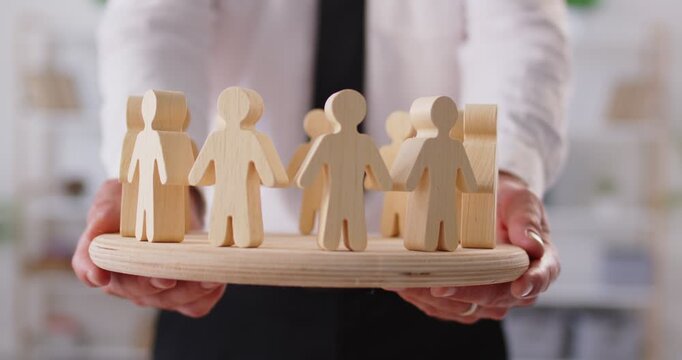 Close up cropped shot of man in shirt and tie reaching out wooden figures of people standing in circle on wooden board forward. Cooperation, teamwork, human resources, community, blockchain concept.