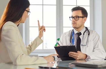 Obraz premium Serious man doctor writing down notes in clipboard while listening to complaints of female patient, giving advice about disease treatment, giving prescription. Health care concept.