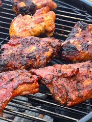 Several portions of marinated pork ribs and chops are cooking on a hot outdoor barbecue grill, featuring a rich, caramelized red-brown glaze and char marks.