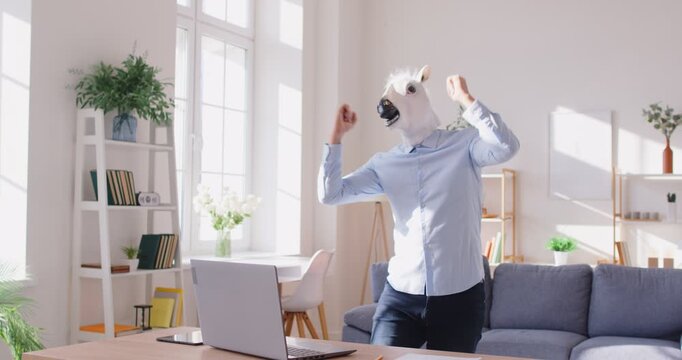 Successful businessman wearing horse mask clenching fists as sign of triumph, posing near workplace with laptop computer, celebrating nice deal with partner, working distantly from home