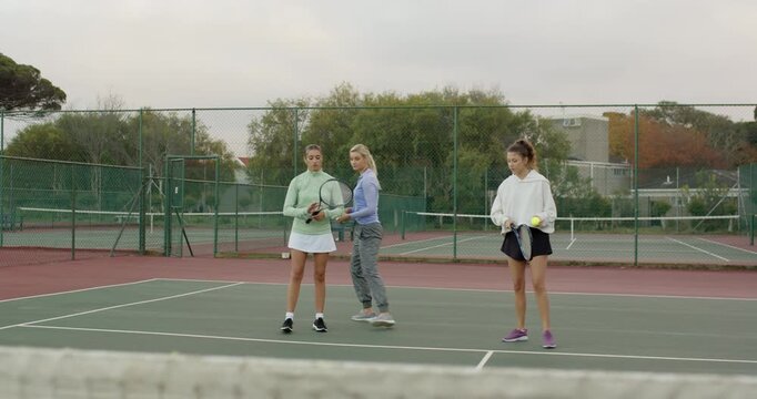 Coach demonstrating forehand, tossing balls as diverse students practicing forehands on hard court