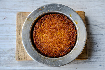 Top view of traditional Turkish künefe dessert with crispy golden kadayıf layers served hot in a metal plate on rustic wooden board.