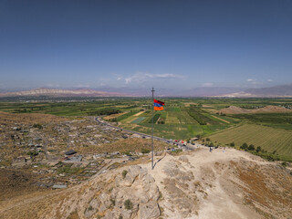 Aerial view of the Armenian flag fluttering boldly atop a hill, overlooking the lush green fields and distant mountains, Khor Virap, Ararat Province, Armenia.