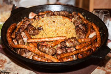 Assorted grilled meat with sausages, minced rolls, and rice pilaf served in a large cast iron pan on a table.