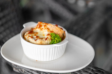 French onion soup in white ramekin, topped with golden melted cheese and toasted bread, garnished with parsley, served on white plate, close-up.