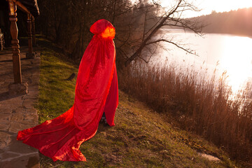 A mysterious figure in a long red hooded cloak stands near a lake at sunset, creating a fairytale...