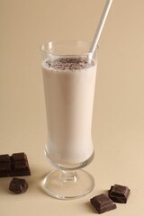 Tasty milkshake in glass and pieces of chocolate on beige background, closeup