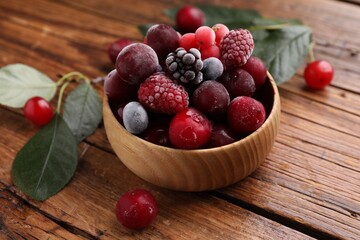 Mix of ripe frozen berries in bowl and green leaves on wooden table, closeup