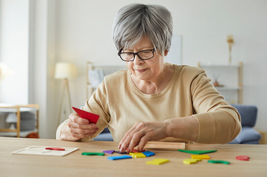 Senior woman sitting at table with colorful wooden pieces, playing memory game at home. Focused pensioner solving tangram puzzle, training cognitive skills for brain health and mental stimulation.