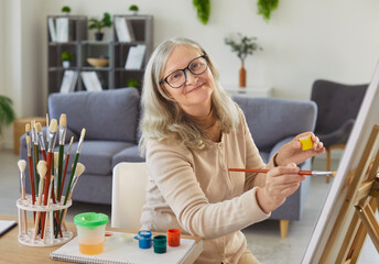 Happy senior older woman painting on easel, holding brush, applying paint to canvas, old lady...