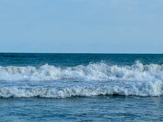 Powerful ocean waves crash onto a sunlit shore beneath a clear sky.