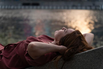 Young woman with red hair in a burgundy dress relaxing by the riverbank at sunset, with warm golden...
