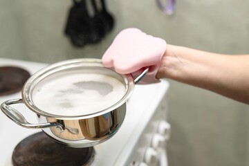 woman's hand holds a small metal pot with boiling water using a special silicone pot holder for safety