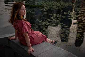 Woman in a burgundy dress sits on a stone embankment, gazing thoughtfully at the river surface with...