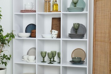 Shelving unit with different dishware, glasses and other kitchen utensils near white wall indoors