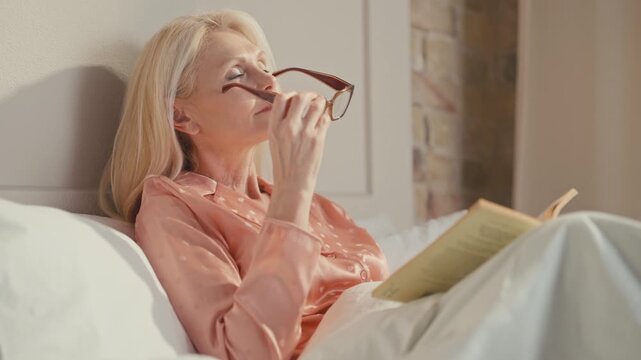 Mature woman with long blonde hair sitting in bed adjusting eyeglasses. Holding open book while wearing peach nightwear. Relaxing indoors, enjoying calm reading time in comfortable bedroom setting.
