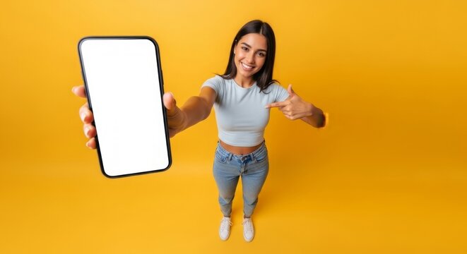 Cheerful young woman in casual clothes points to a large smartphone with a blank white screen, viewed from a high angle against a bright yellow background.