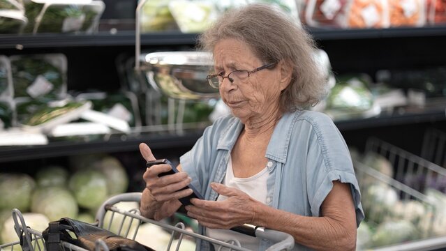 An elderly senior woman is grocery shopping in a store, examining her phone while pushing a cart filled with food items in the produce section. - Powered by Adobe