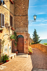 Fototapeta premium Tuscany, Italy, Pienza historic center street with stone houses, wooden door, climbing plants, terracotta pots, lantern, and scenic panoramic view