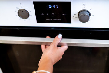 A close-up of a woman's hand, lightly touching the stainless steel handle of a modern oven with a...