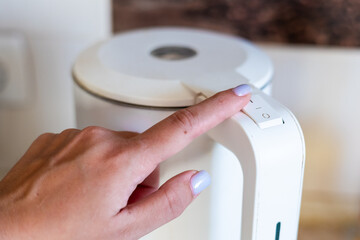 A close-up of a hand pressing the switch on a modern white kettle in a bright kitchen setting.
