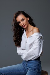 Obraz premium Beautiful tall thin brunette with curly hair in the studio, cyclorama. The girl is standing in jeans and a white shirt, gray background. Studio photo shoot for a model. Portrait. Beautiful makeup