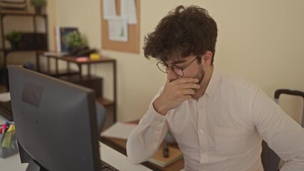 Young man with beard working at computer in office room, looking thoughtful and engaged, surrounded by books and plants, creating a casual yet professional setting indoors. - Powered by Adobe