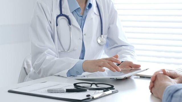 Female doctor reviewing health information on digital tablet and explaining diagnosis to patient during clinical consultation in medical office. Medicine and healthcare concept