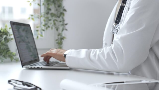 Male doctor wearing a white coat and stethoscope is typing on a laptop in a bright medical office, showcasing the intersection of healthcare and technology, close up. Medicine and health care concept