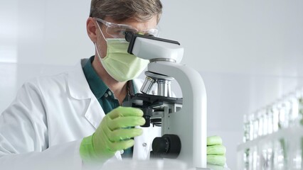 Adult male scientific professional analyzing sample through microscope, wearing green protective gear in sterile laboratory setting. Medicine, healthcare and science concept