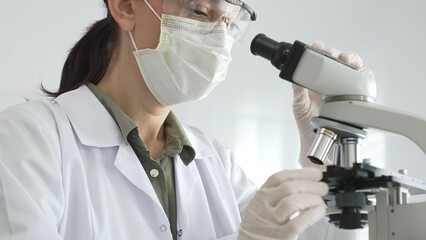 Female scientist examining microscope slide, wearing white protective gear while conducting medical...