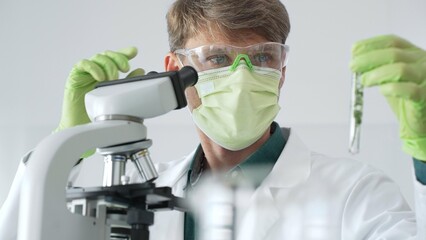 Adult male scientist wearing green mask, gloves and goggles using microscope and analyzing sample...
