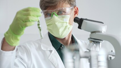 Adult male scientist wearing green mask, gloves and goggles using microscope and analyzing sample...