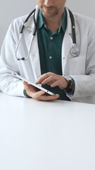 Doctor man wearing lab coat over green shirt, and stethoscope, is using digital tablet in modern...