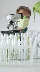 Man scientist wearing green protective gloves and mask is looking at green plant in test tube near...