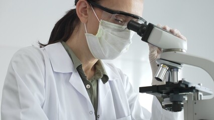 Female scientist examining microscope slide, wearing protective gear while conducting medical...